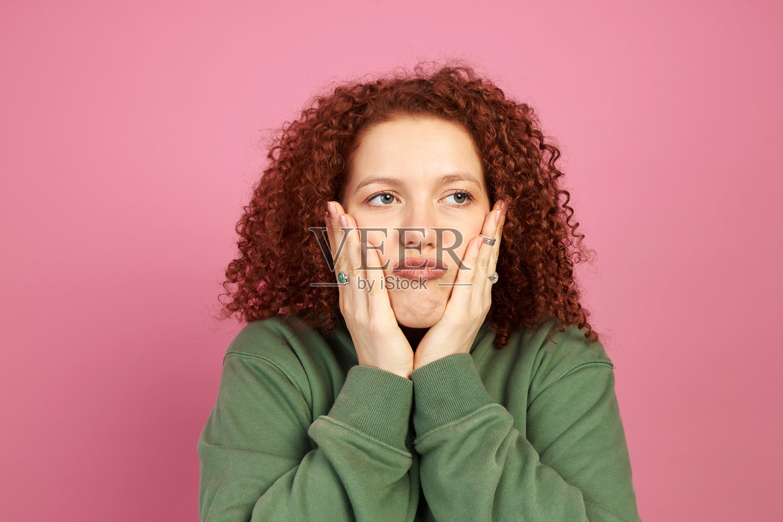 漂亮的无聊搞笑的红发千禧女孩，卷发可爱，双手托着脸颊，撅着嘴唇，侧目凝视，背景为粉色。面部表情与情感。照片摄影图片