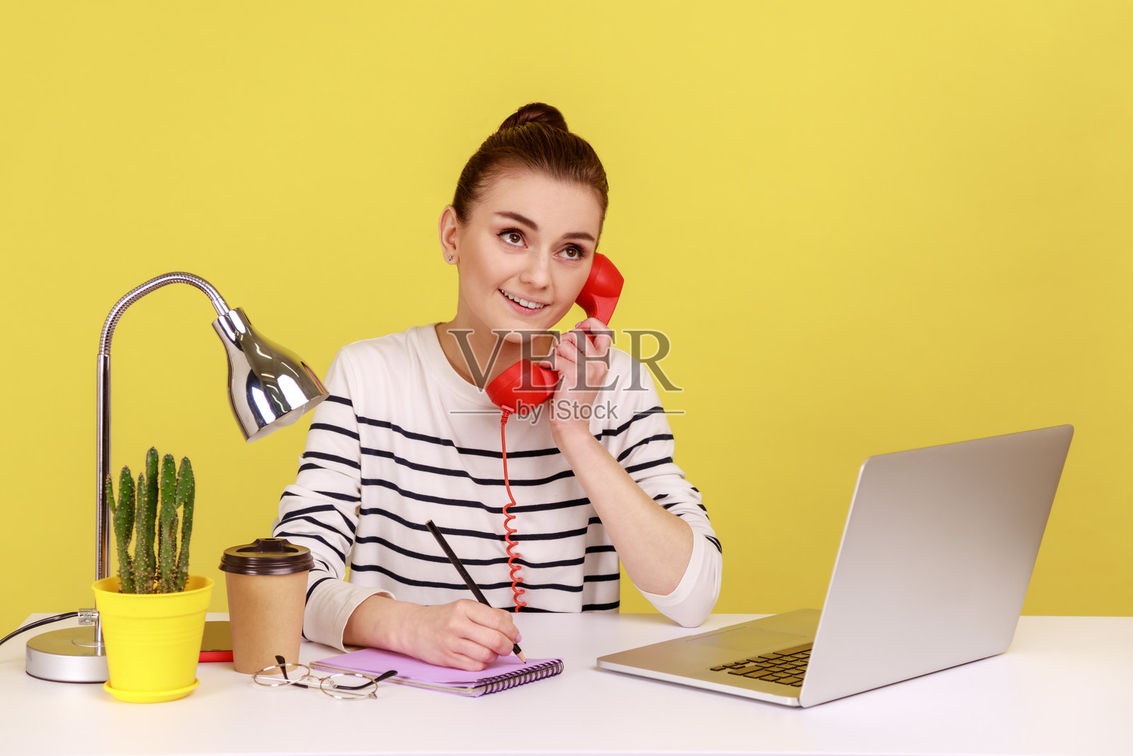 女人坐在工作场所，手持红色复古电话，面带梦幻般的表情，正在记笔记本上写字。照片摄影图片