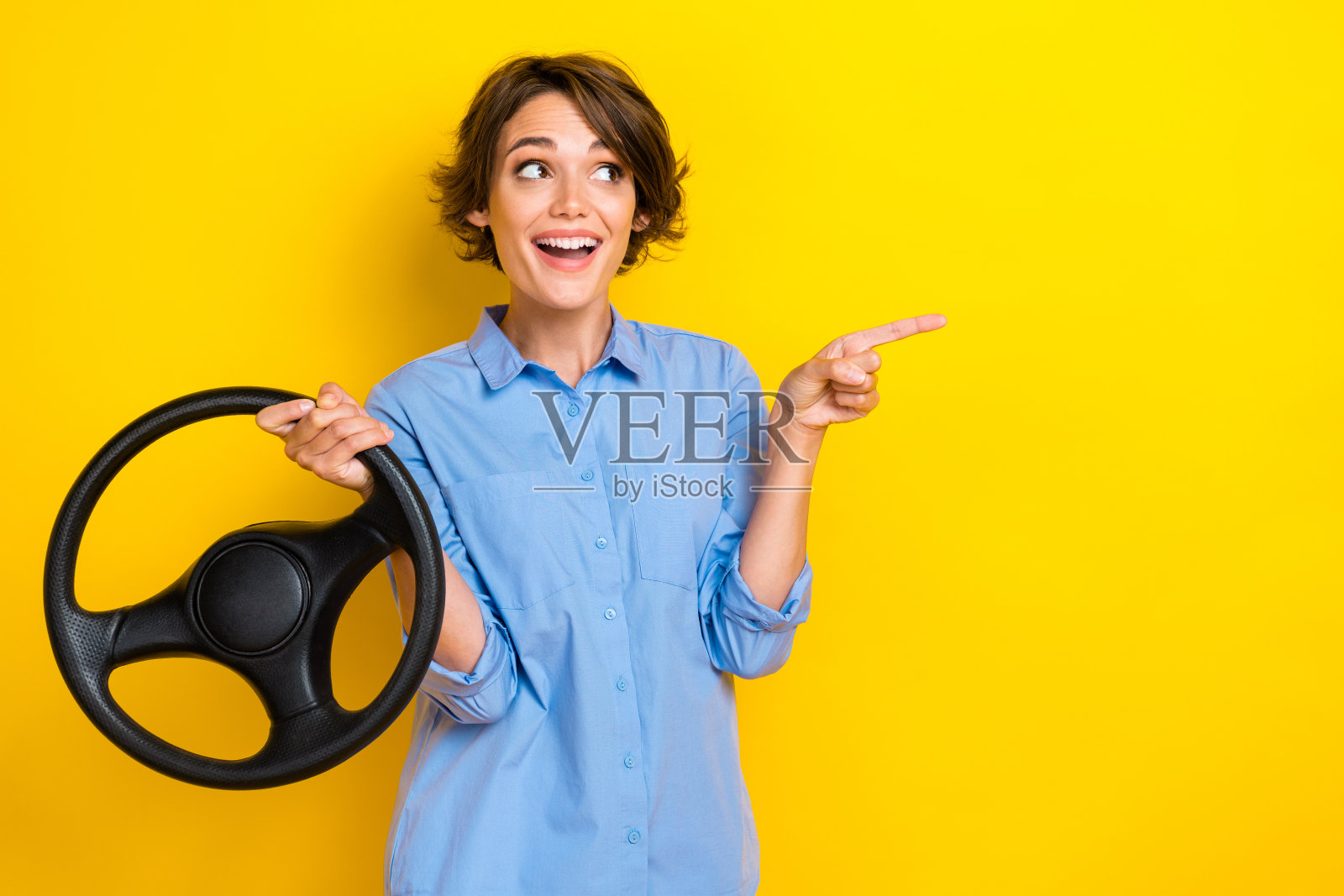 穿着蓝色衬衫的可爱兴奋女孩指着空白处，骑在汽车上，背景为黄色。照片摄影图片