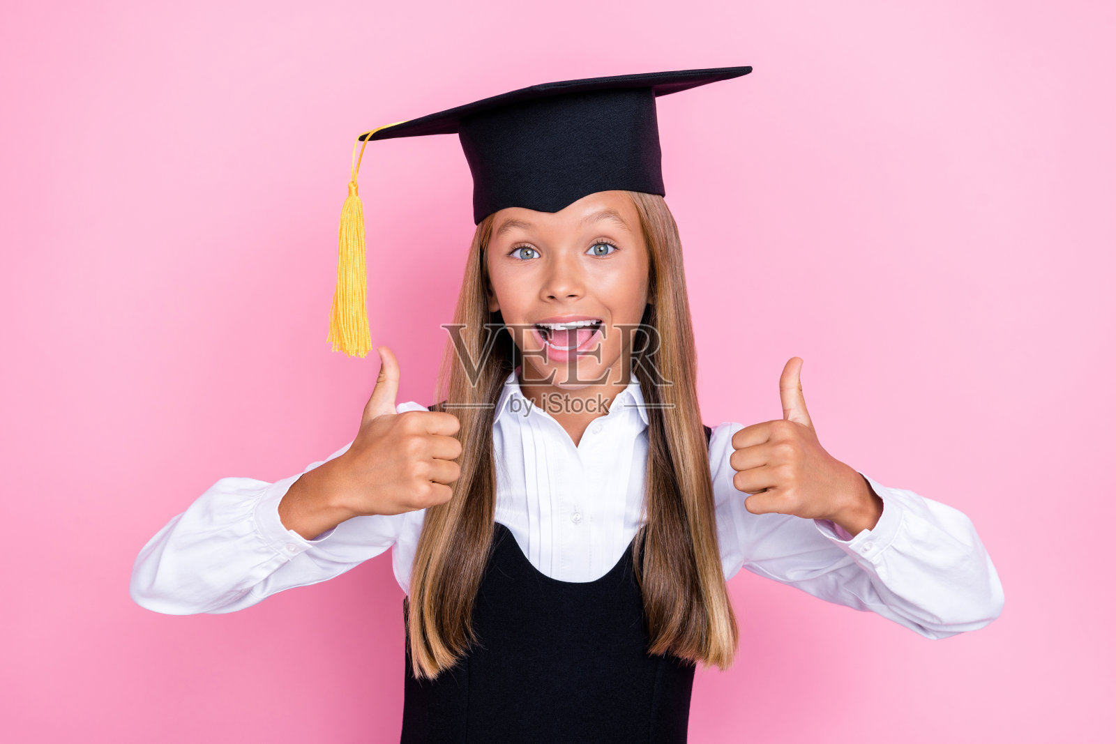年轻可爱的女孩穿着校服和帽子，兴奋而积极，心情愉快，竖起大拇指，背景为粉色。照片摄影图片