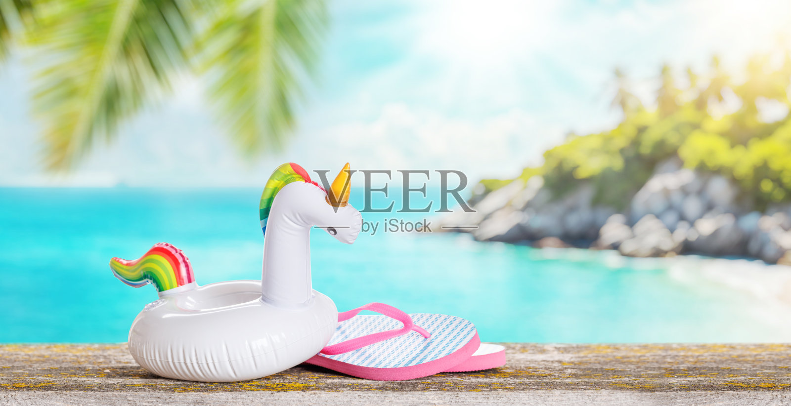 桌子上放着人字拖和一个漂浮的火烈鸟，背景是阳光明媚的大海。照片摄影图片
