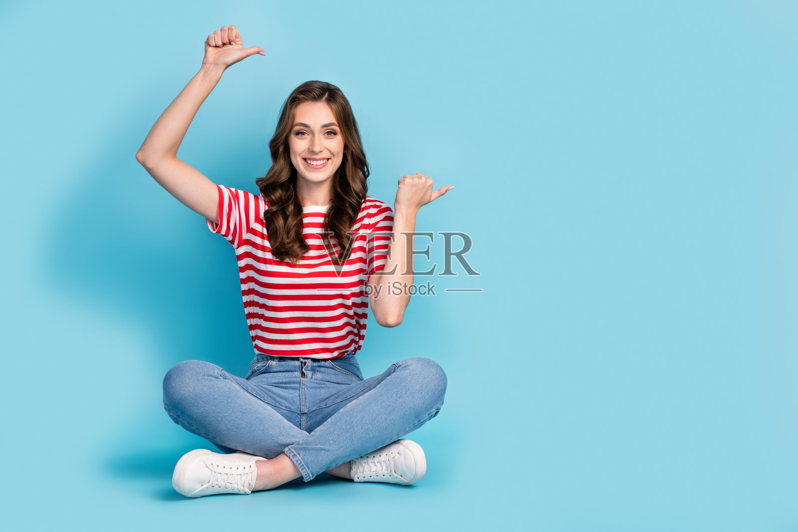 可爱的漂亮女士穿着条纹T恤，双手竖起大拇指指向空白空间，背景为蓝色。照片摄影图片