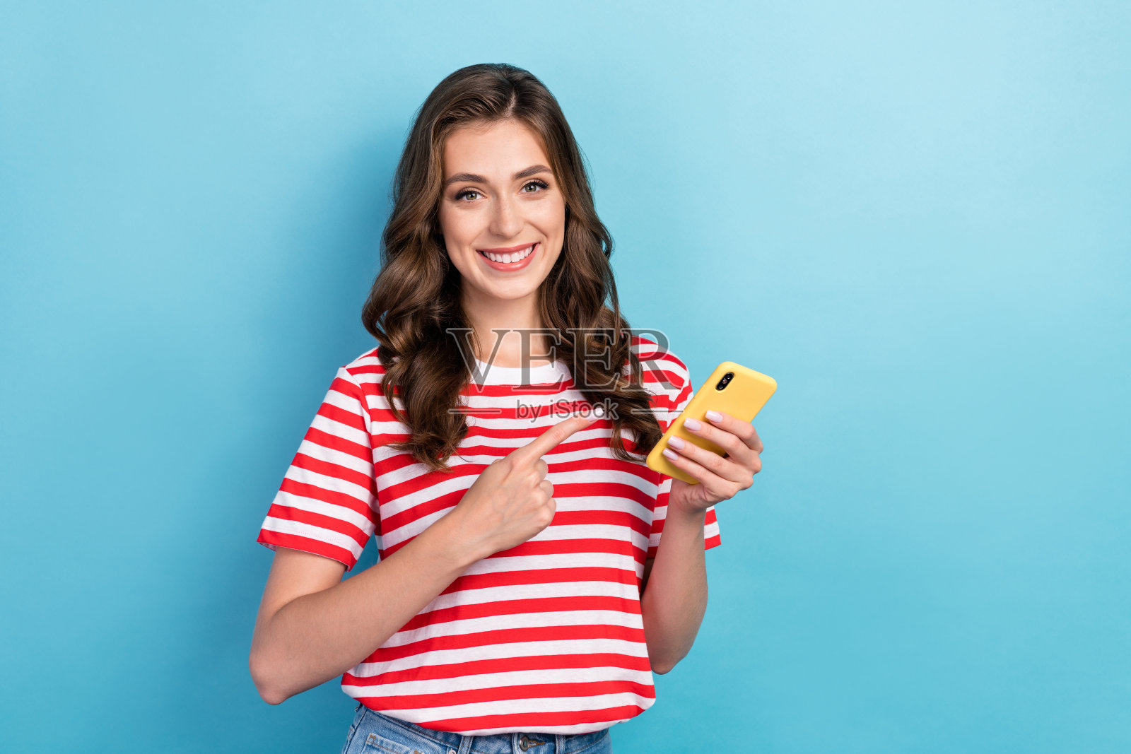 年轻迷人漂亮的女孩用手指指向她的新智能手机，低价高性能，背景为蓝色。照片摄影图片