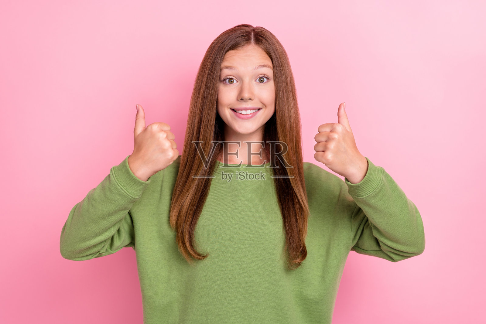 可爱的乐观友好的女孩，直发，穿着绿色毛衣，竖起大拇指，背景为粉色。照片摄影图片