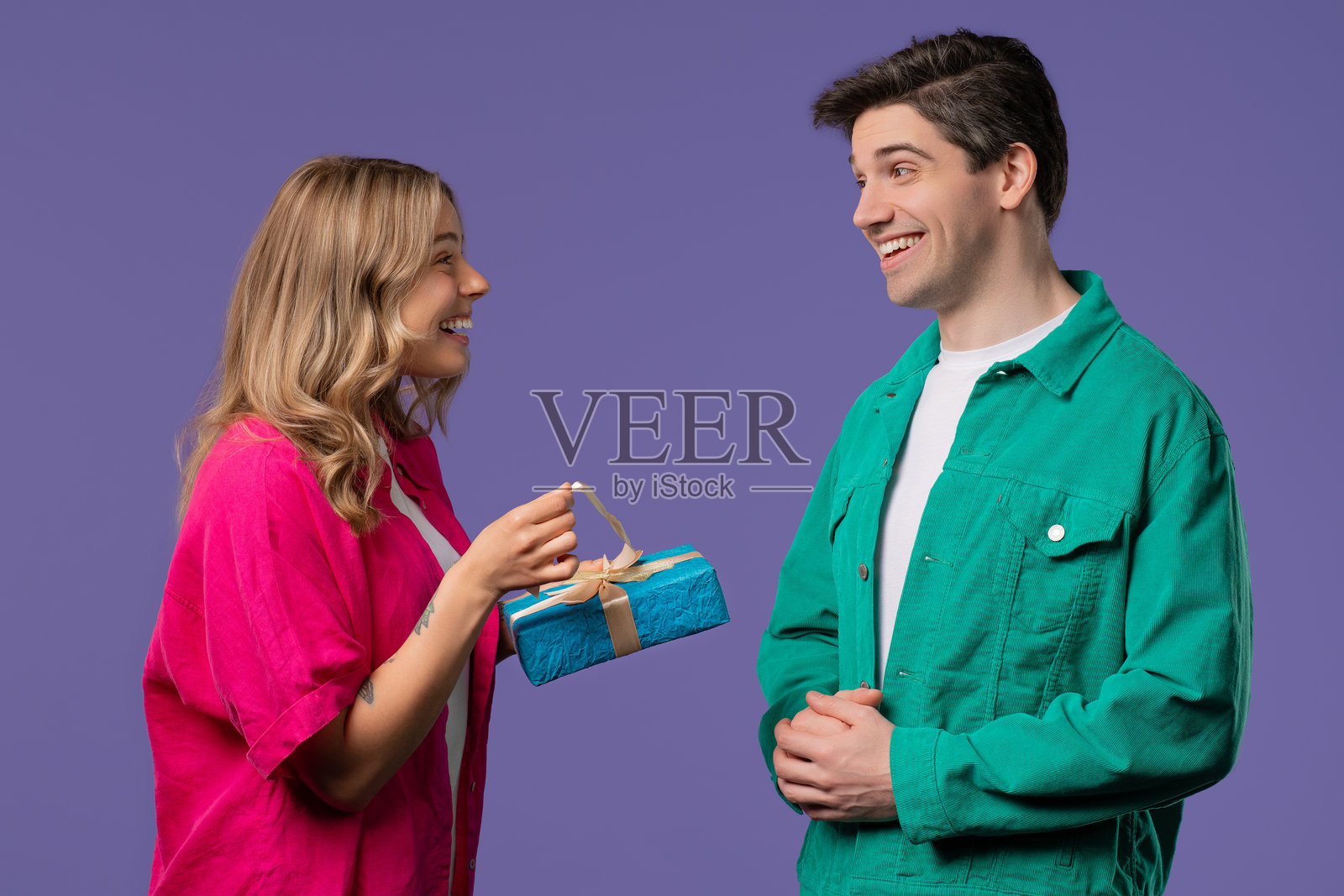 年轻的美国情侣在紫色的摄影棚背景下。白人男性向妻子或女友赠送带蝴蝶结的礼物。生日、庆祝、惊喜、周年纪念的概念。照片摄影图片