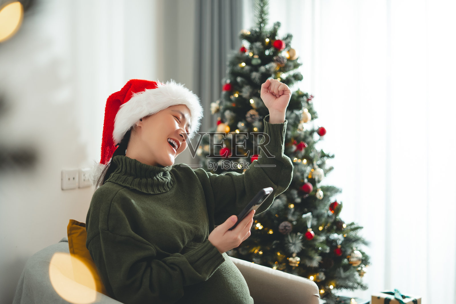 戴着圣诞帽的亚洲孕妇在装饰有圣诞树的客厅里使用智能手机，开心兴奋并做出胜利手势。照片摄影图片