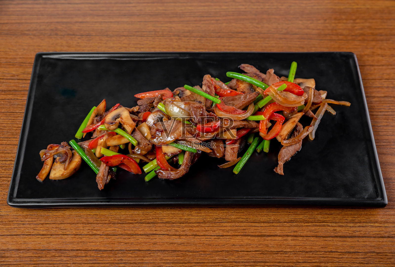 黑色盘子上放着炸猪肉和牛肉，配有大蒜箭、蘑菇和蔬菜，摆放在木桌上。照片摄影图片