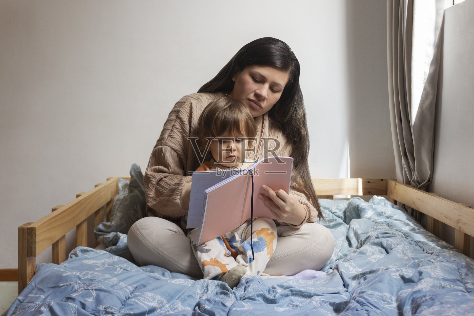 母亲抱着一岁大的宝宝坐在床上，她正在给他讲故事，母子共度美好时光。照片摄影图片