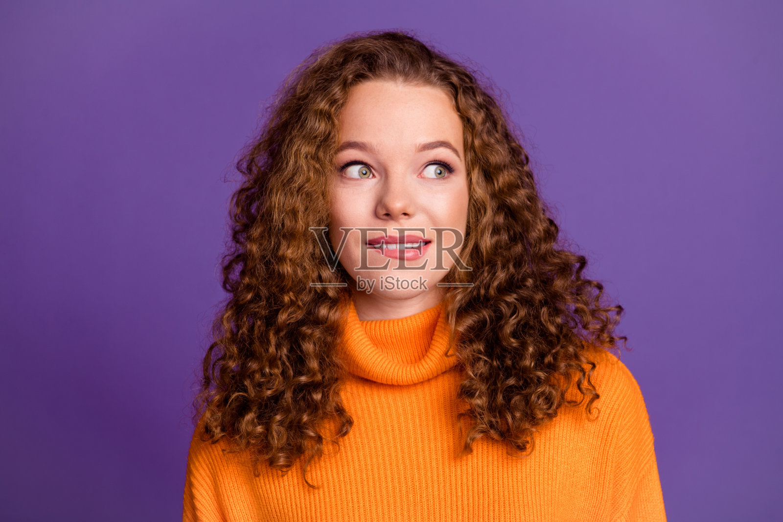 穿着橙色时尚高领毛衣的年轻女性，波浪形红发，咬着嘴唇，兴奋地思考，心情愉悦，背景为紫色。照片摄影图片