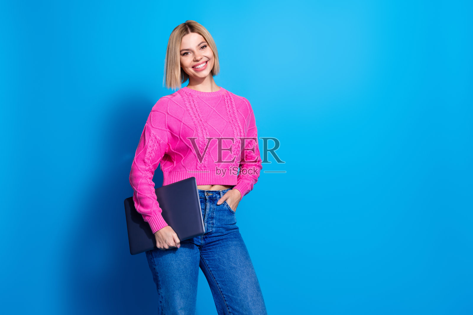 穿着粉色毛衣的漂亮年轻女性在蓝色背景前使用空白笔记本电脑的照片照片摄影图片