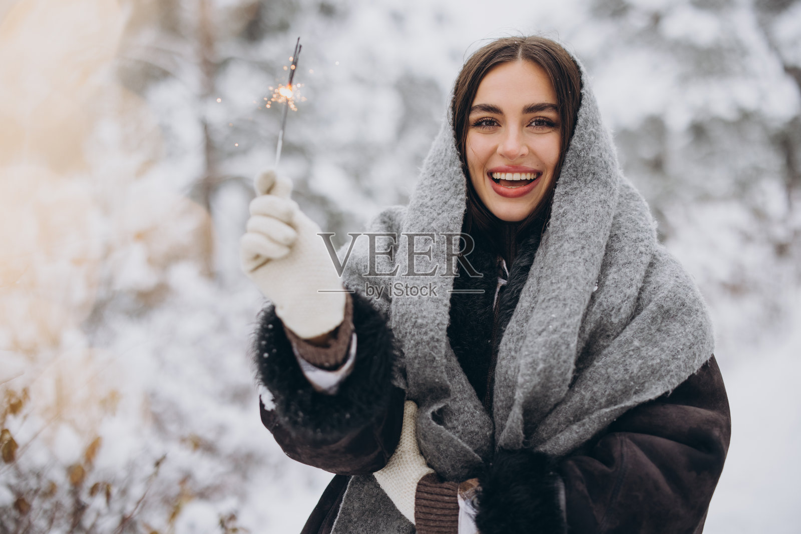 在雪-covered森林中，快乐的漂亮女人穿着针织围巾和手套，手持烟花并摆姿势。照片摄影图片