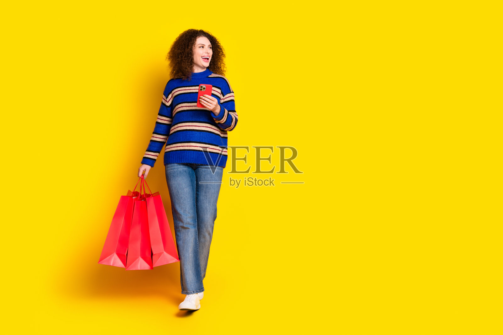 穿着毛衣的积极女性全身照，手提购物袋，手持智能手机，望向空旷的空间，背景为黄色。照片摄影图片