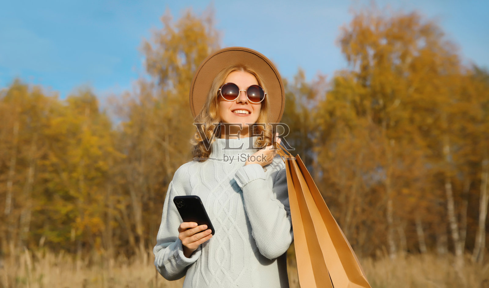 时尚快乐的年轻女性在秋季公园里，手持购物袋，微笑着使用手机。照片摄影图片