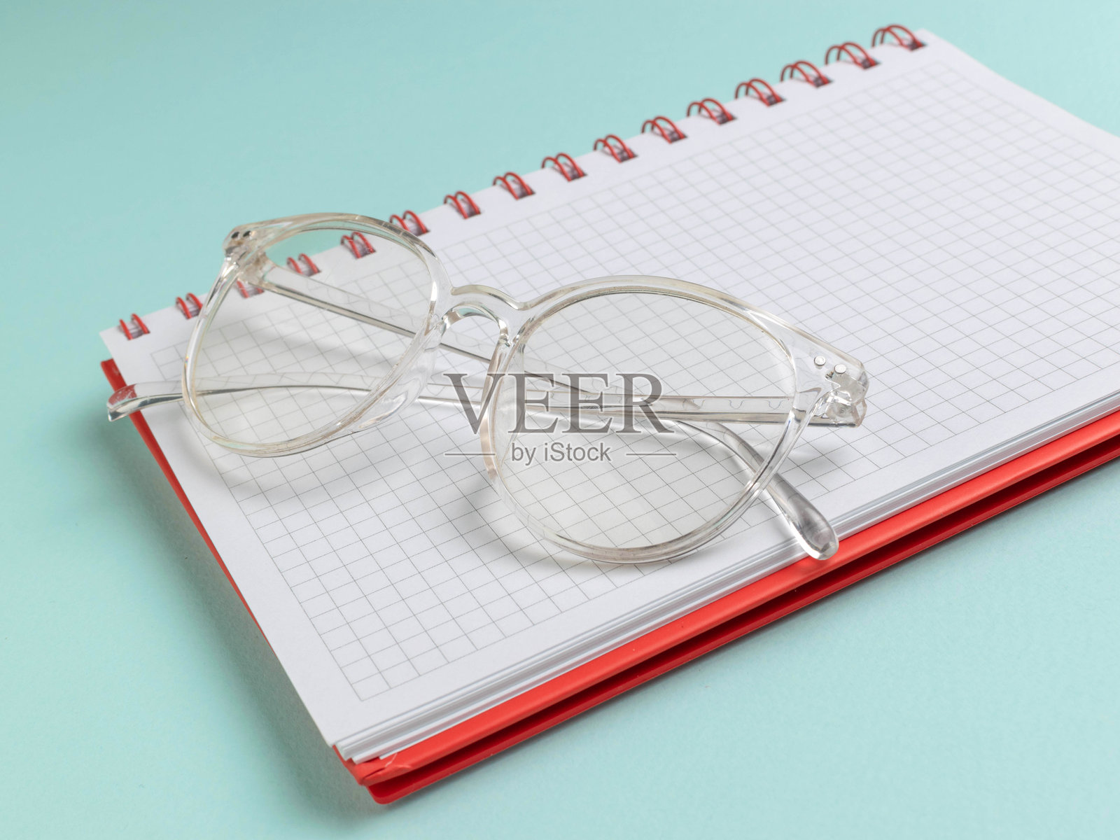 清晰的眼镜放在红色螺旋笔记本上，笔记本上有方格纸，背景为绿色。照片摄影图片