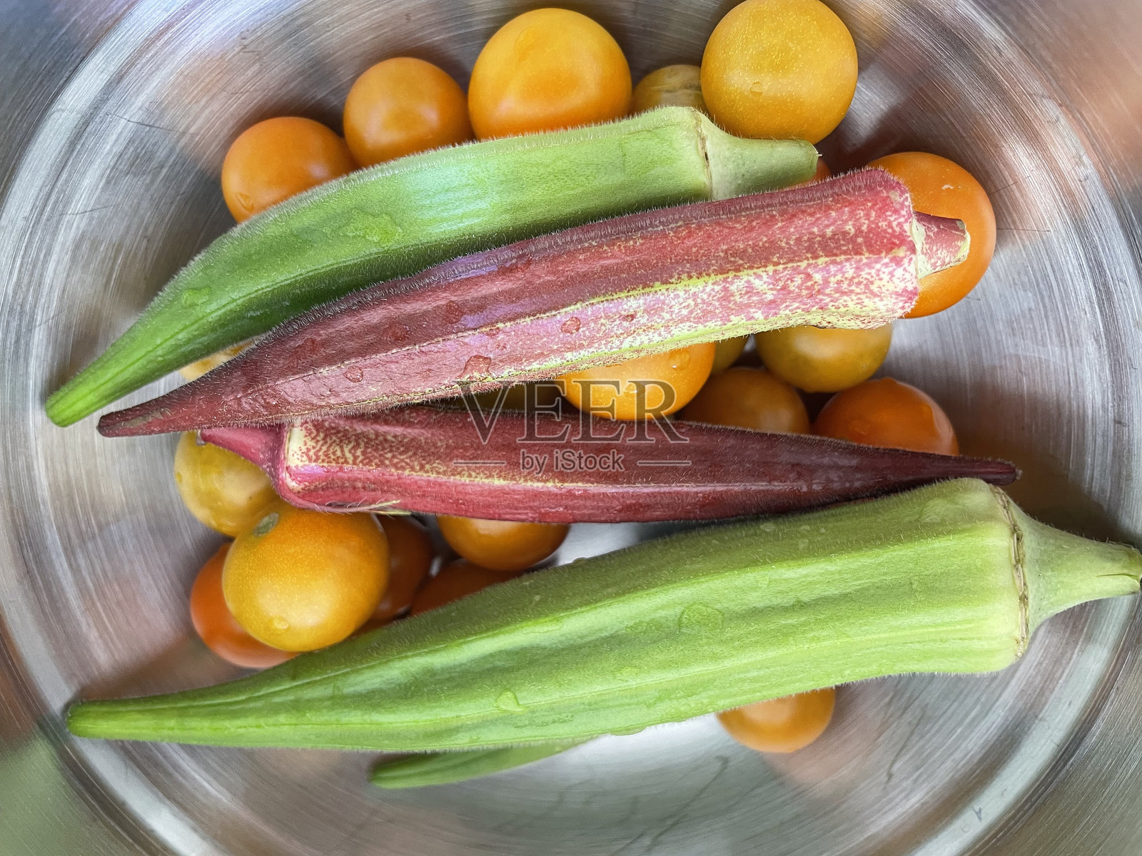 钢锅中垂直放置的秋葵荚与橙色迷你番茄特写，带有水珠。照片摄影图片