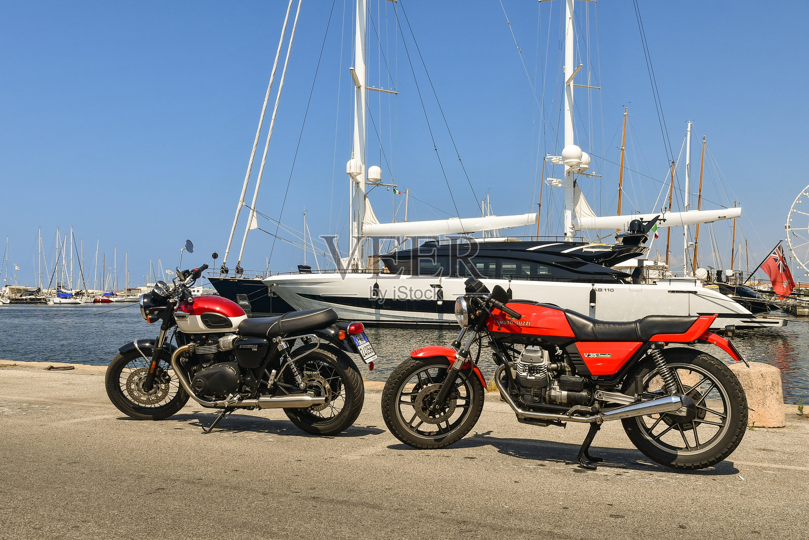一辆凯旋博纳维尔T100和一辆摩托古兹V35伊莫拉摩托车停在码头上，背景是豪华游艇，位于意大利托斯卡纳卢卡省的海滨小镇维亚雷焦。照片摄影图片