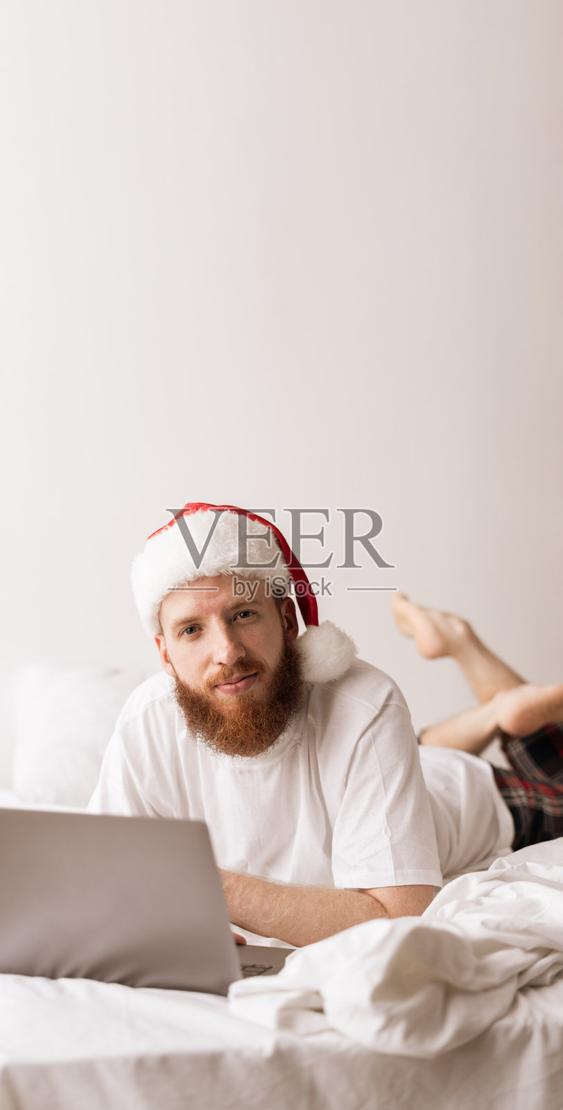 戴着圣诞帽的胡子男人在圣诞早晨从床上远程工作照片摄影图片