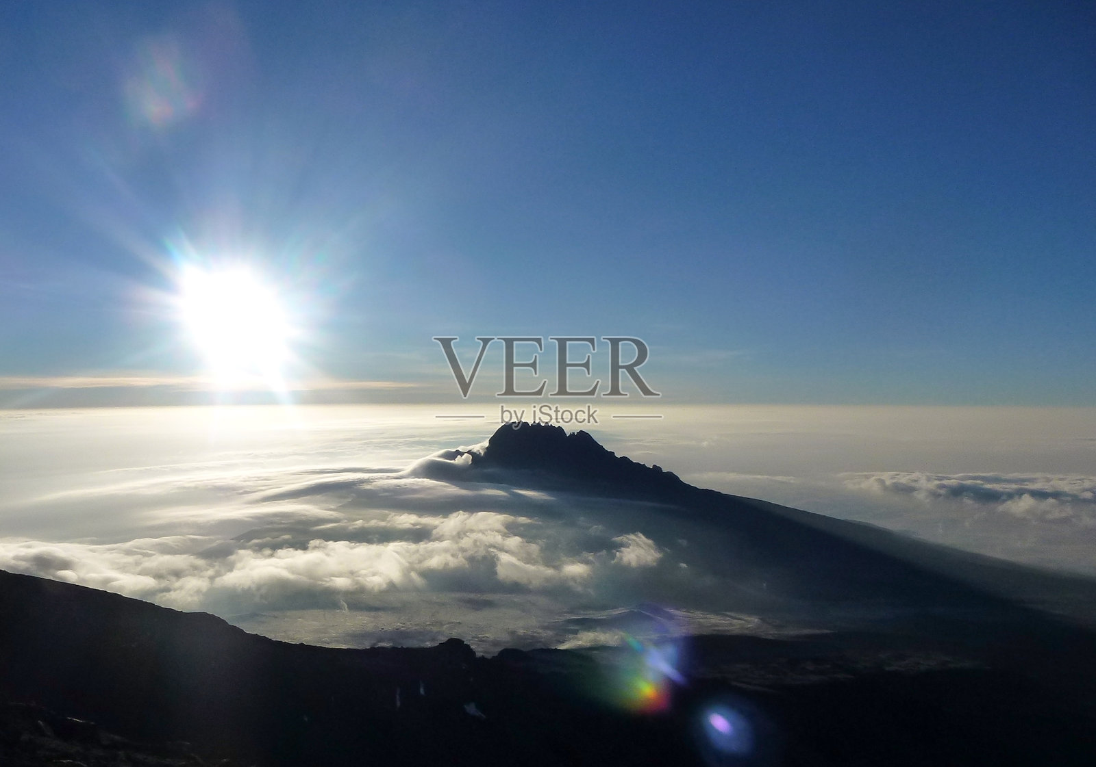 从上方俯瞰一座附近的山峰，耸立在云层之上，背景是明亮的阳光和蓝天。照片摄影图片