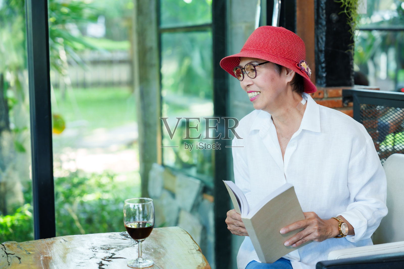 穿着白衬衫和红色帽子的亚洲老年女性在现代餐厅里微笑着阅读一本书。木桌上放着一杯酒。周围有自然光，大窗户外面是绿色植物。照片摄影图片