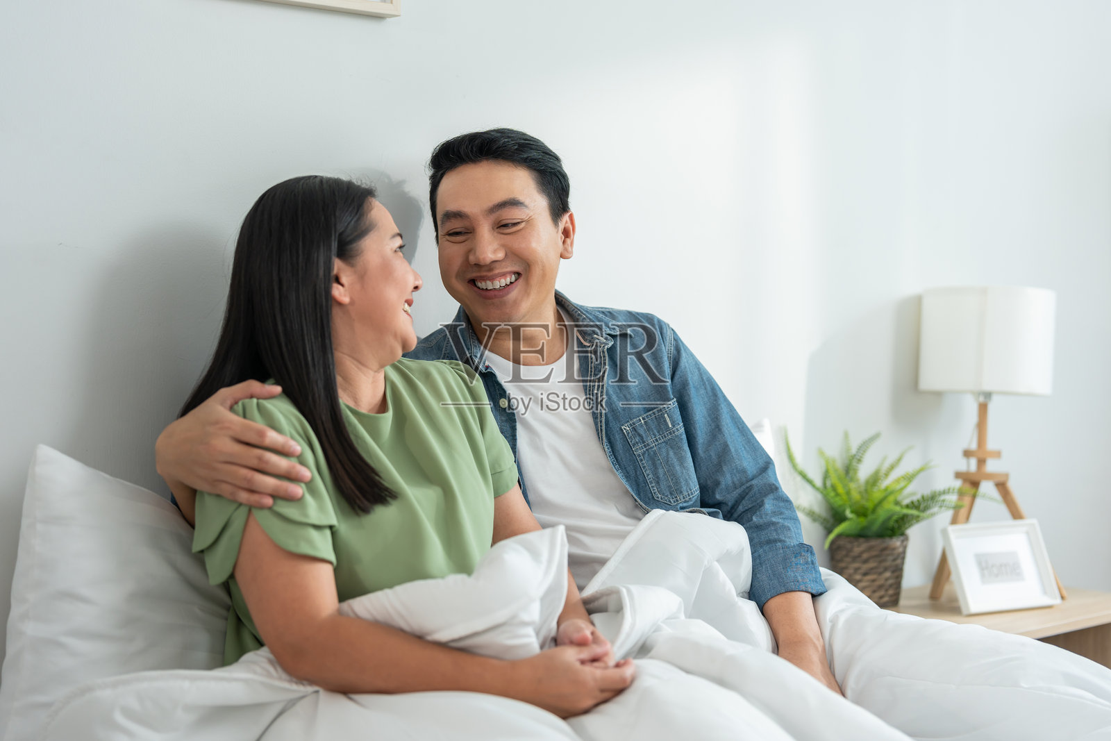 亚洲成熟夫妇在家中床上醒来后坐着。迷人的男女感到快乐和放松，享受彼此交谈，度过早晨的闲暇时光。照片摄影图片