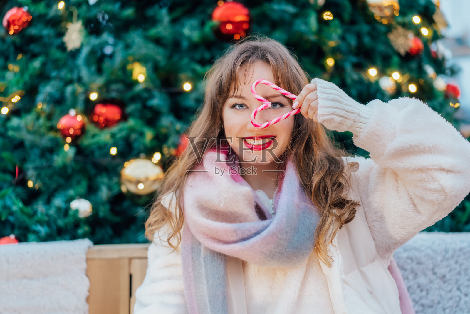 年轻微笑的女性透过用糖果手杖做成的心形观看，背景是装饰好的圣诞树。快乐的新年庆祝圣诞节假期。分享爱与慈善的时刻。照片摄影图片