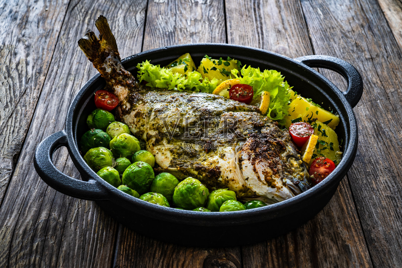 黑色盘子里盛着烤海鲷、煮土豆和布鲁塞尔芽菜，摆放在木桌上。照片摄影图片