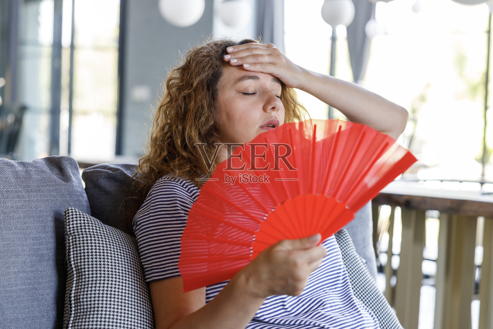 年轻女性在家沙发上挥舞扇子，因过热而感到不适。照片摄影图片