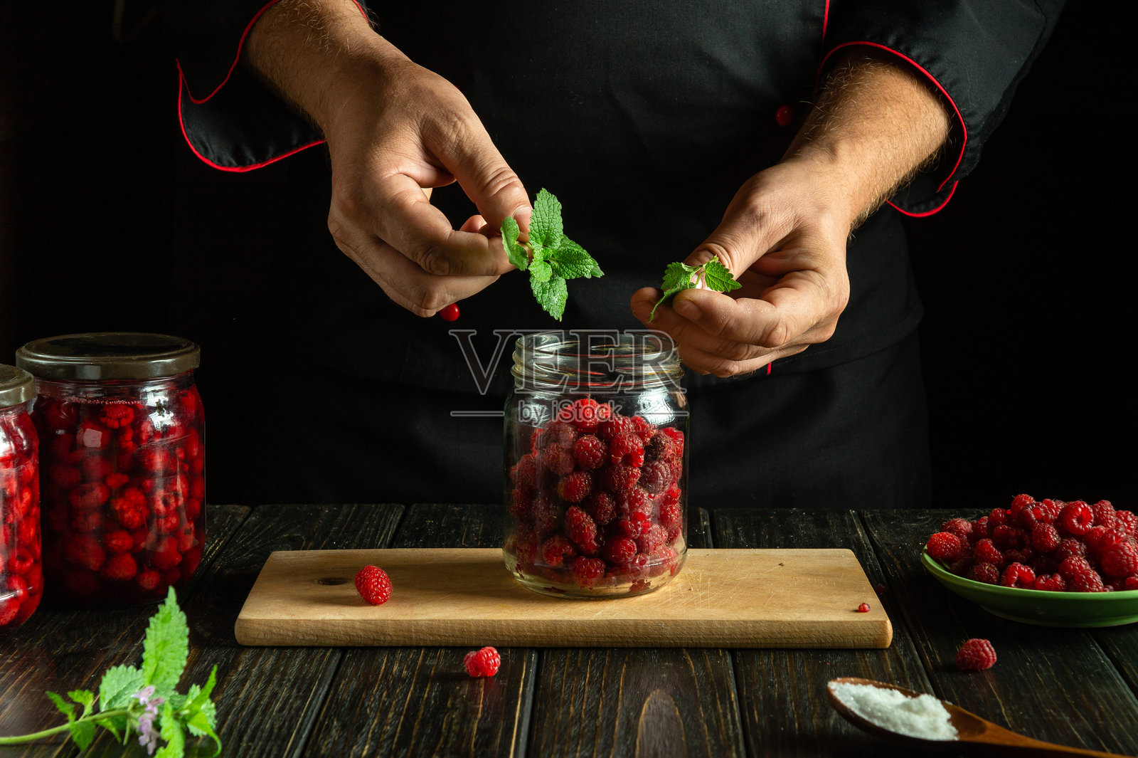 厨师在罐子里腌制红覆盆子。厨房桌上制作甜饮料的概念。将薄荷叶加入覆盆子中。照片摄影图片