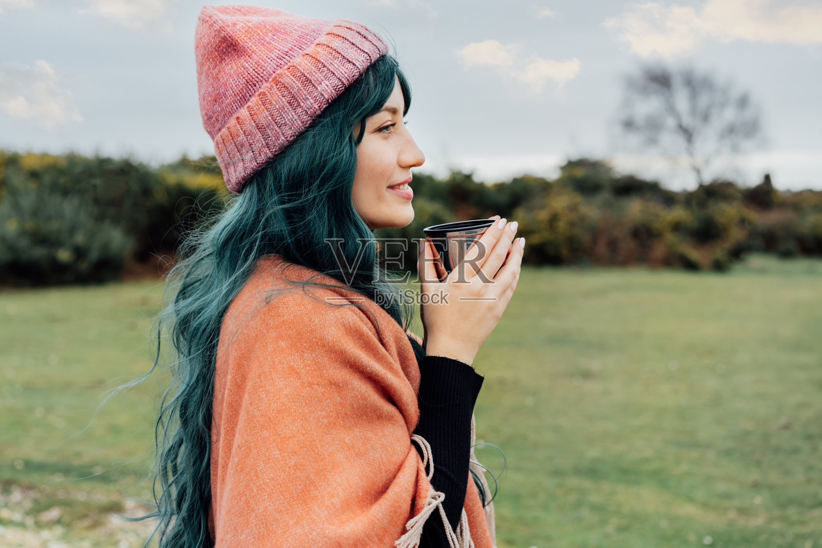 戴着帽子的微笑女性，裹着披肩，手持保温杯饮用热饮，享受秋季风景。感受和谐、放松与个人满足。当地旅行生活方式。照片摄影图片