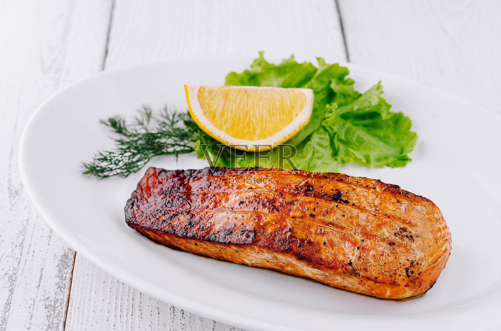 白盘上放着烤三文鱼排，旁边有柠檬和生菜。照片摄影图片