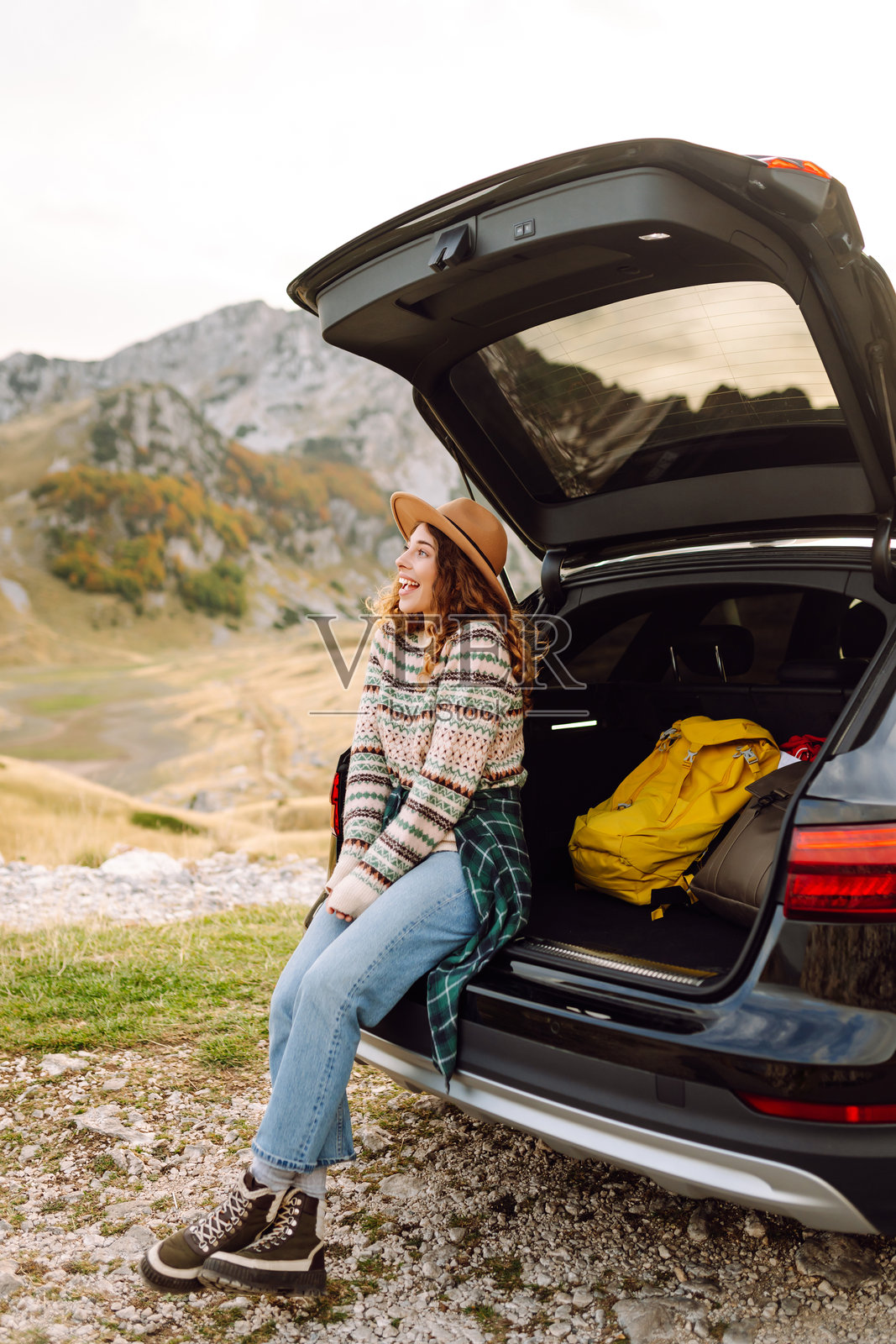 一位卷发戴帽的女旅行者微笑着，悠闲地坐在她的黑色SUV后面。照片摄影图片