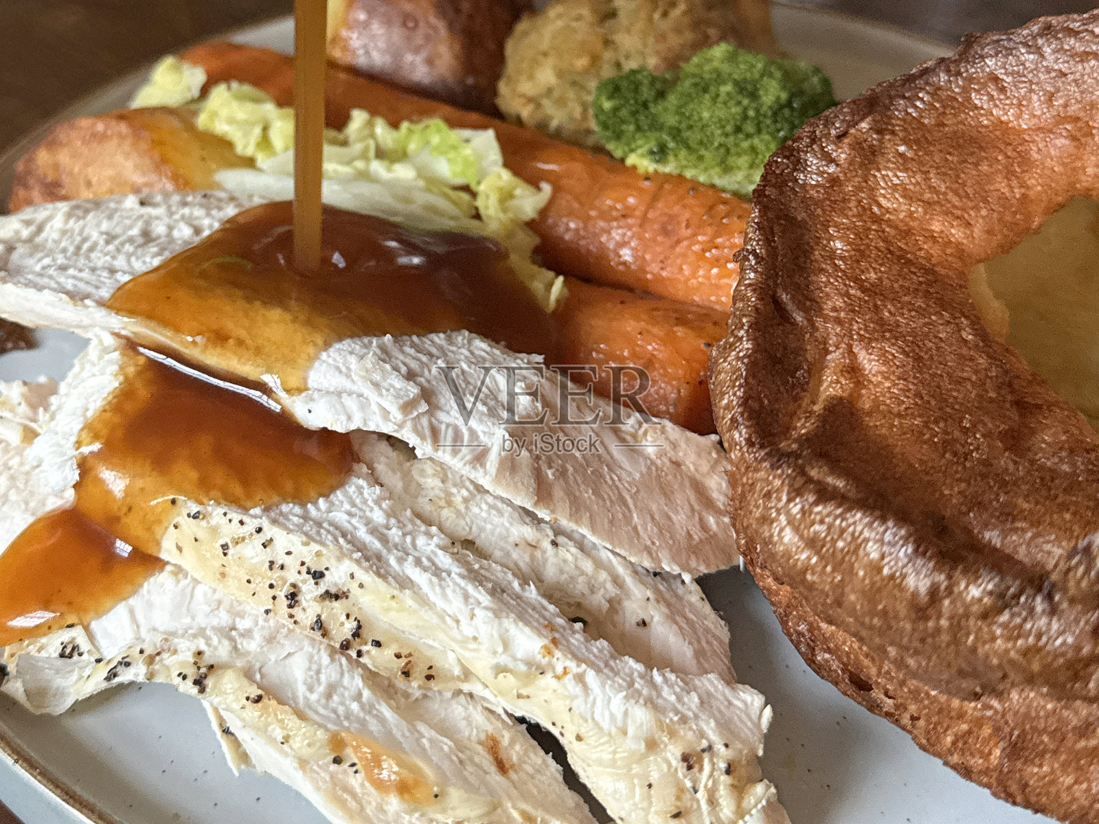 全景图像，肉汁倒在白色盘子上，盘中包含周日烤肉晚餐，约克郡布丁、胡萝卜、卷心菜、烤土豆、西兰花、鸡胸肉片、鼠尾草洋葱面包填料球，俯视角度，重点在前景。照片摄影图片