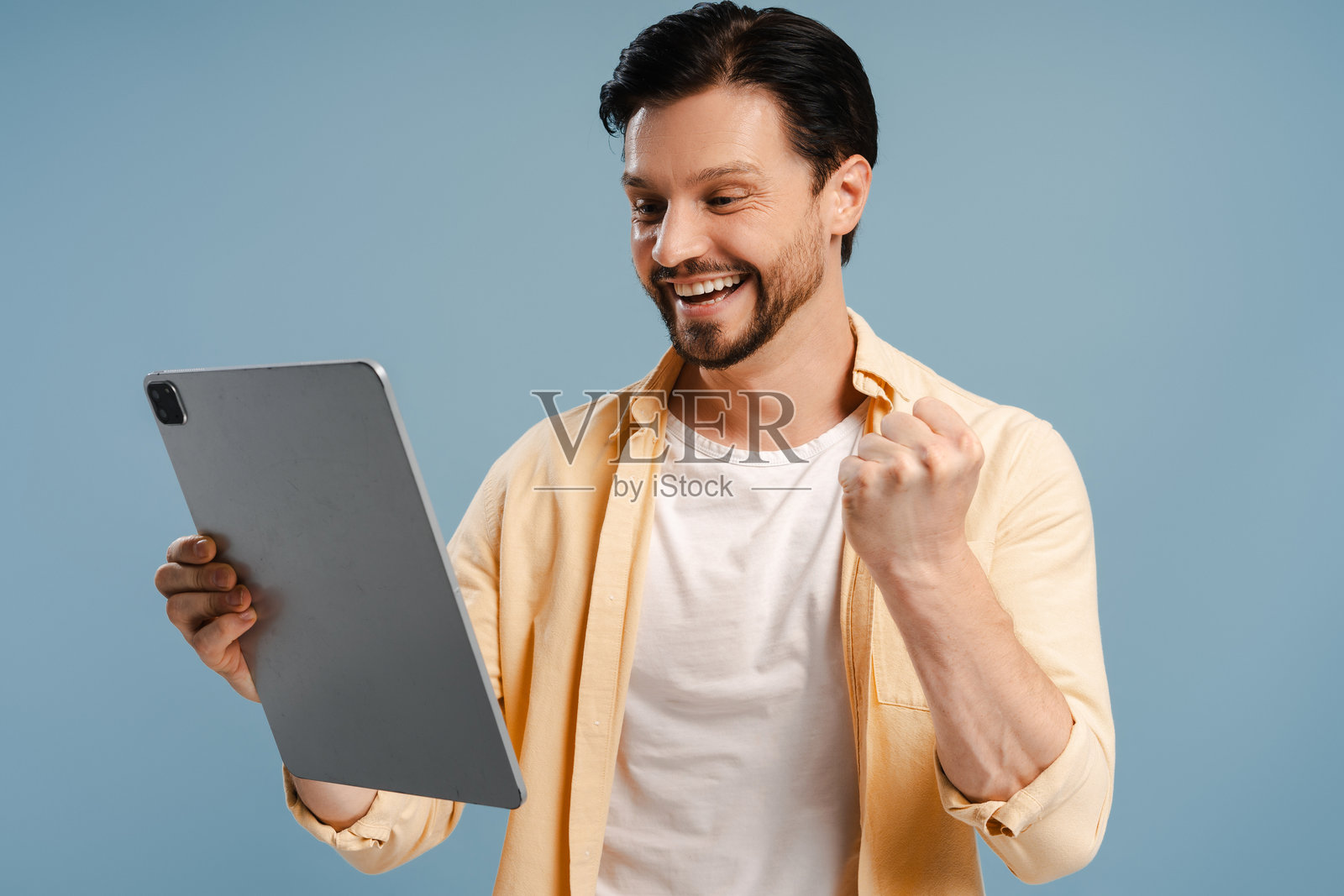 兴奋的络腮胡男子拿着数位平板电脑庆祝成功，背景为蓝色照片摄影图片