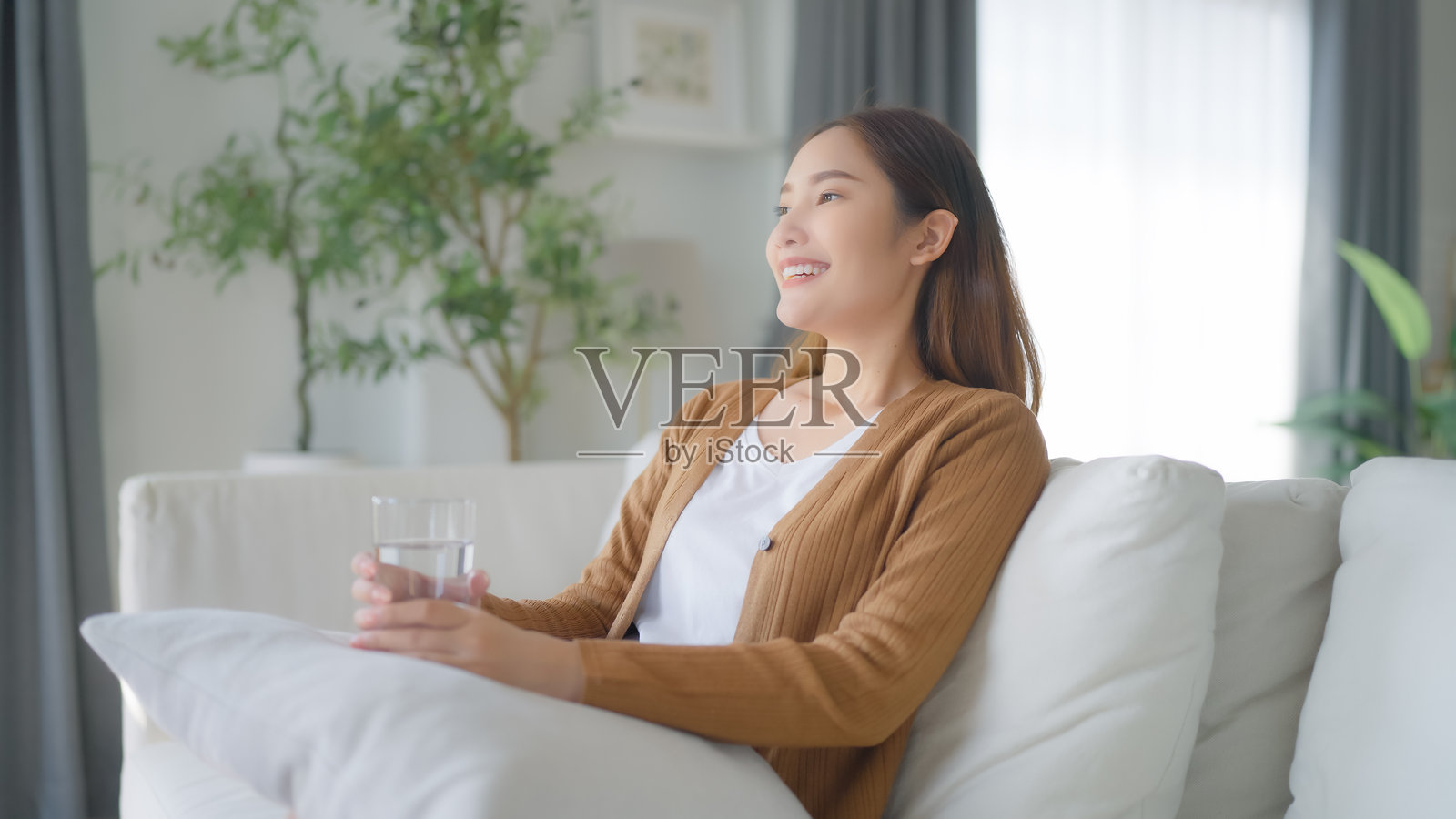 快乐的年轻亚洲女性坐在客厅的沙发上喝水照片摄影图片