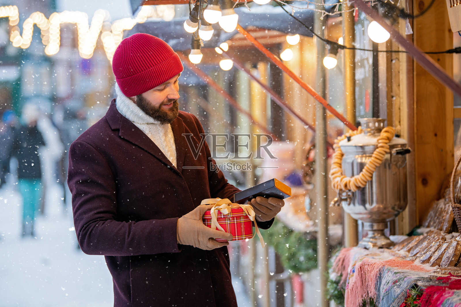 快乐微笑的男子穿着冬季服装，在雪天的市中心广场的圣诞市场上挑选和购买礼物。照片摄影图片