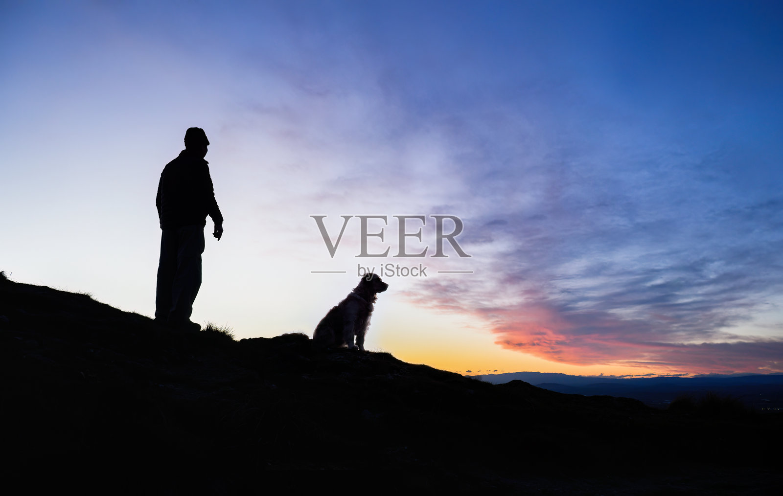 日落时分，男人和他的狗在山顶上的剪影图像。特马塔峰，霍克斯湾。照片摄影图片