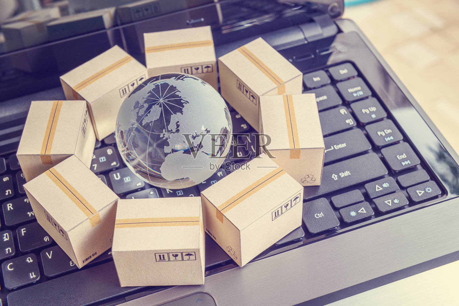 国际货运或在线购物电商概念：纸箱围绕着一个带有世界地图的透明水晶地球，放置在电脑笔记本键盘上。照片摄影图片
