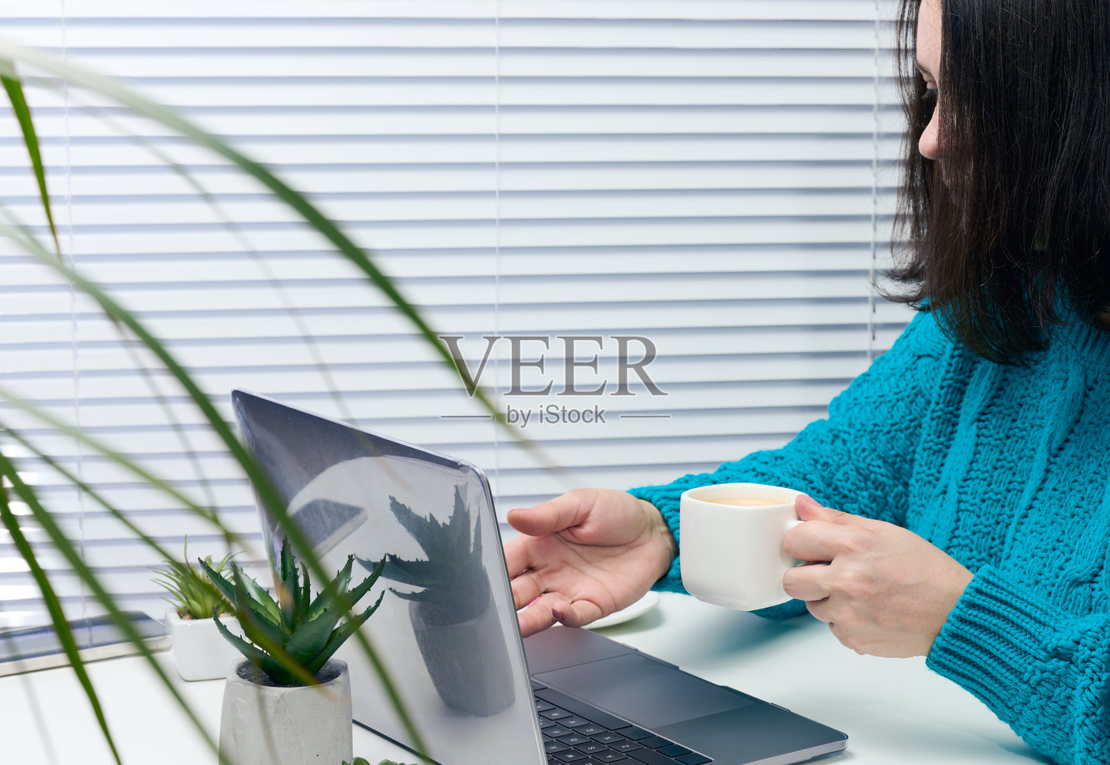 穿着蓝色毛衣的女性坐在桌子前，手中拿着笔记本电脑和一杯咖啡。自由职业者的工作场所，在咖啡馆休息。照片摄影图片