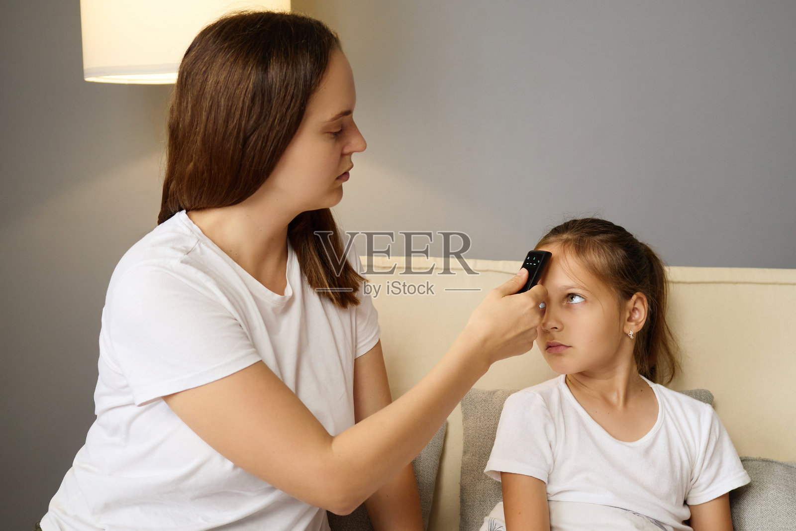 担心的母亲用电子温度计给女儿测量体温，而小女孩在沙发上休息，因流感感到不适，正在家中寻求照顾。照片摄影图片