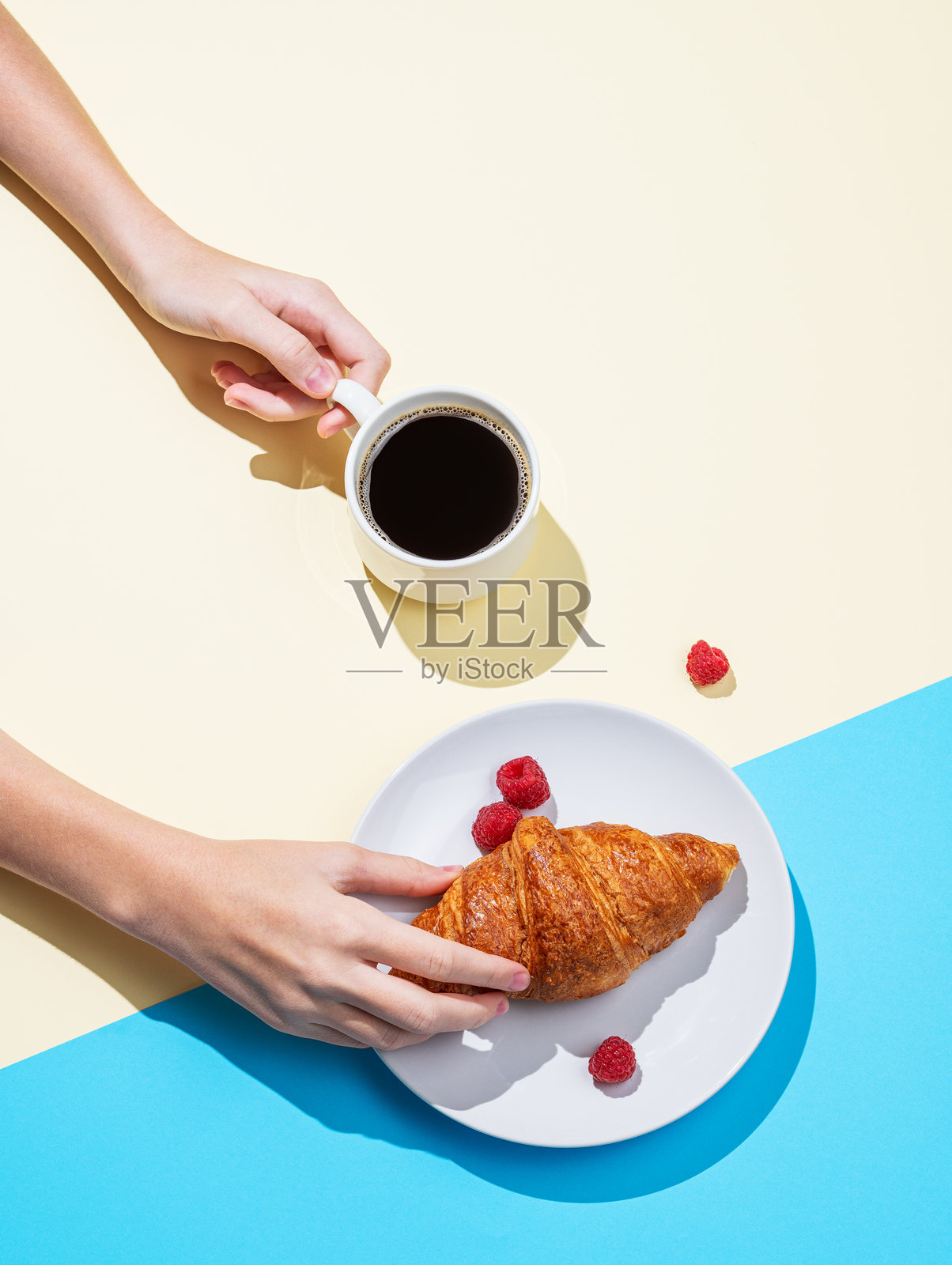 女性手中拿着新鲜的可颂面包和一杯浓缩咖啡，背景为黄色和蓝色。照片摄影图片