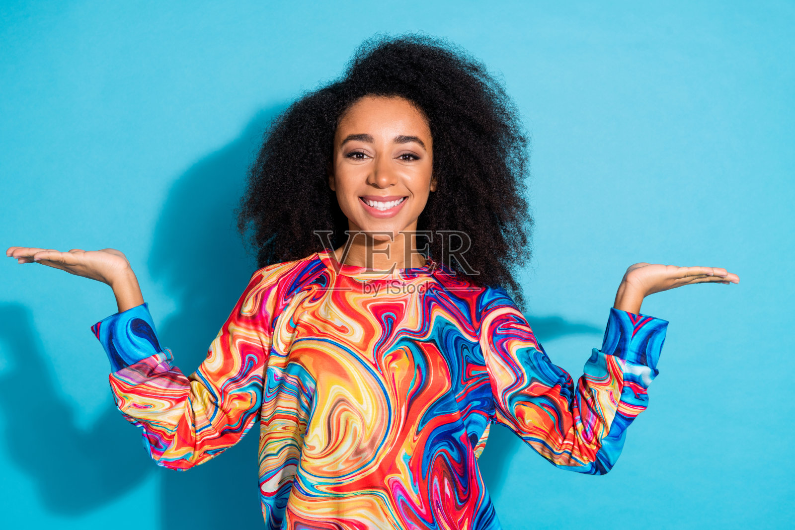 美丽的卷发女性穿着彩色毛衣，双手比较视频，背景为蓝色。照片摄影图片