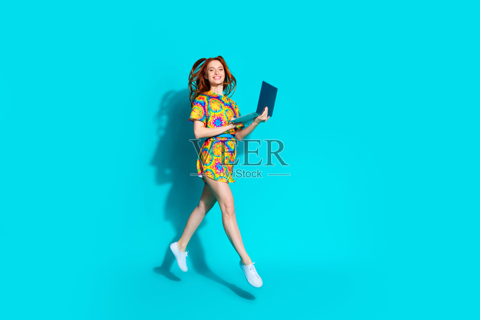 全身照，年轻可爱的女士穿着时尚多彩的服装，跳跃着飞起，手中拿着笔记本电脑，背景为青色。照片摄影图片