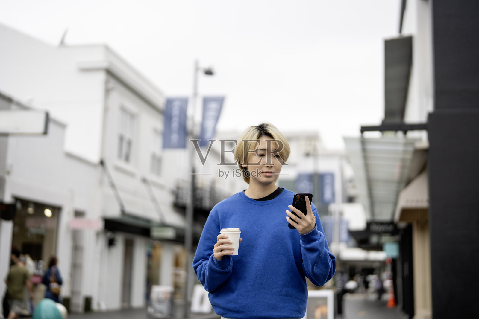 一位时尚的年轻亚洲女性站在狭窄的街道上，查看她的智能手机，手里拿着一杯咖啡。照片摄影图片