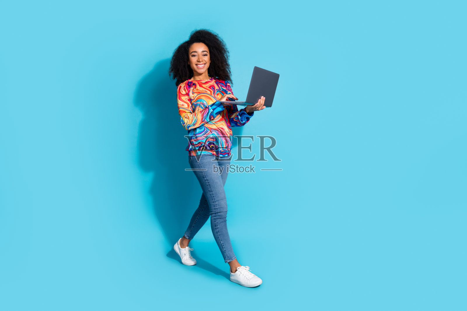 全身照，漂亮的年轻女孩手持笔记本电脑，穿着时尚多彩的服装，背景为蓝色。照片摄影图片