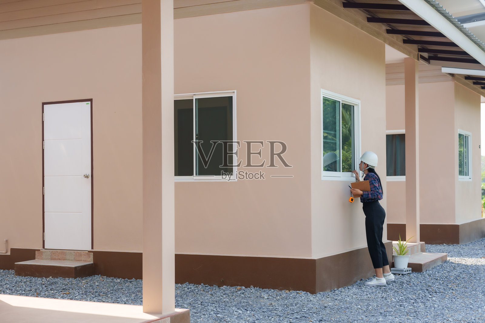 亚洲女性建筑工人戴着安全帽，正在检查房屋外观，手持夹板记录，进行交接过程，检查质量和细节，户外工地，现代房屋，阳光明媚。照片摄影图片