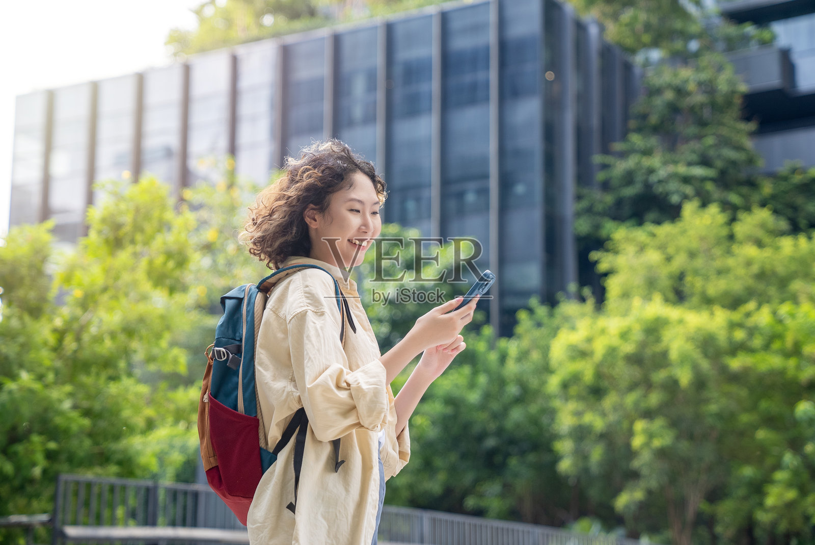 一位年轻女性背着背包，边走边查看智能手机，穿行于现代化的绿意城市景观中。照片摄影图片