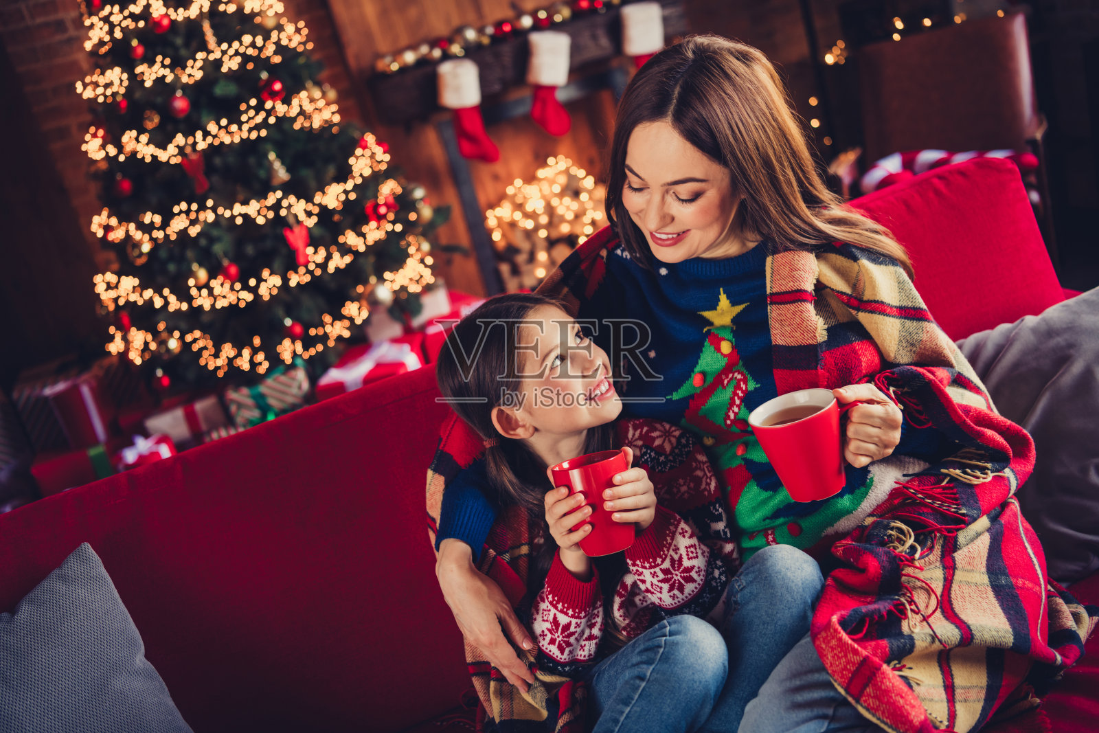 母女两人裹着毯子，拥抱着喝可可，庆祝节日的圣诞时光，室内灯光璀璨。照片摄影图片