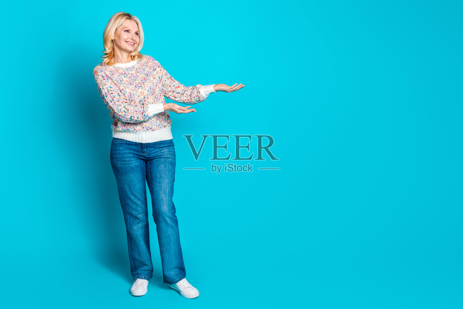 穿着毛衣的优雅老年女性在青色背景前手持空白广告牌的全身照片照片摄影图片