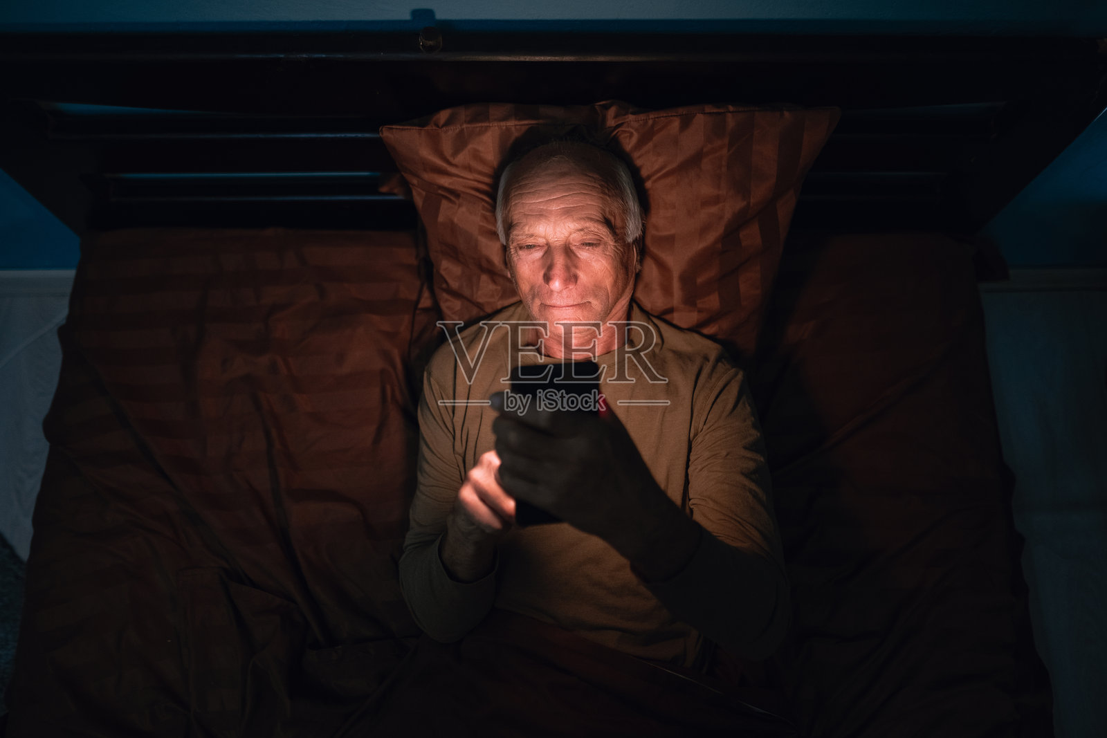 老年男人晚上在卧室躺在床上用手机的俯视图。疲惫的年长男性，无法入睡。失眠和手机成瘾照片摄影图片