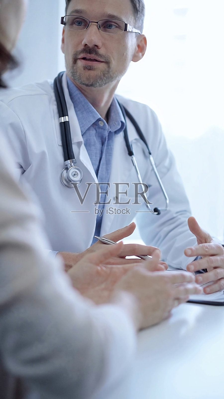 医生和病人：医生穿着白色医用外套和蓝色衬衫，在诊所咨询期间用手势示意，医学概念照片摄影图片